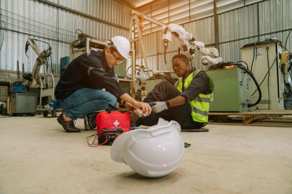 Worker receiving first aid after an injury in an industrial factory setting, with safety gear and equipment visible. Worker receiving first aid after an injury in an industrial factory setting, with safety gear and equipment visible.