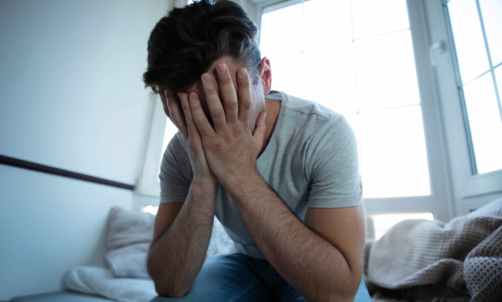 Stressed man sitting on a bed with his face in his hands, showing emotional distress and anxiety.
