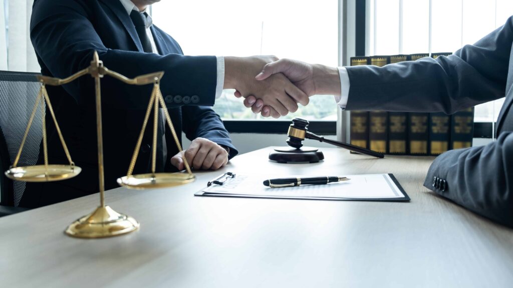 Attorney and client shaking hands in law office with legal documents, gavel, and scales of justice on desk